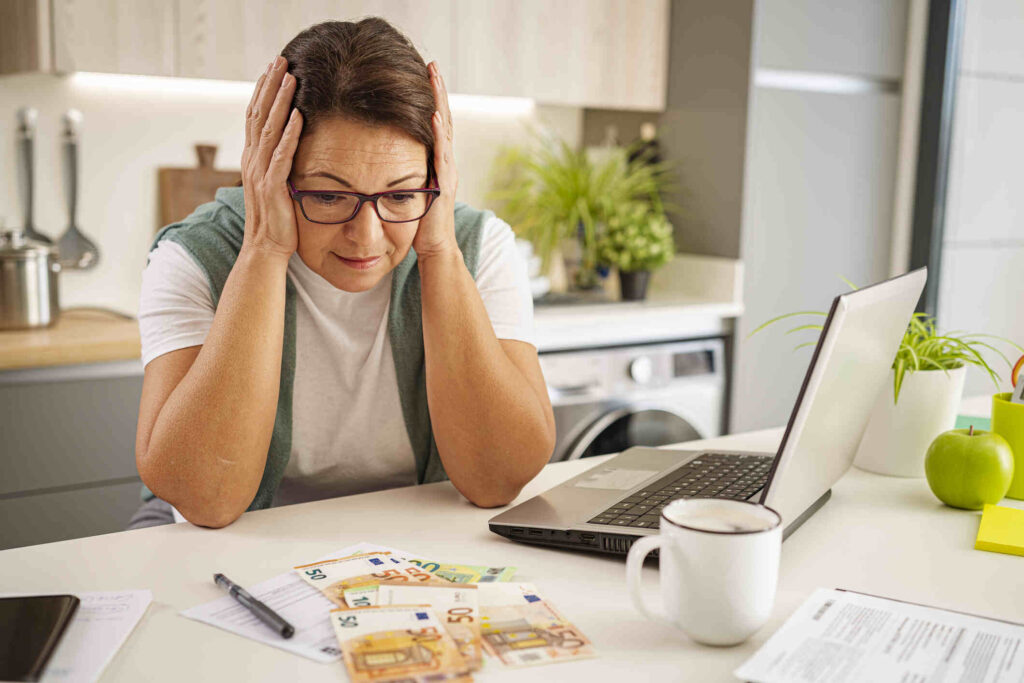 A middle-aged woman with a worried expression on her face sitting at a table, surrounded by bills and financial documents.