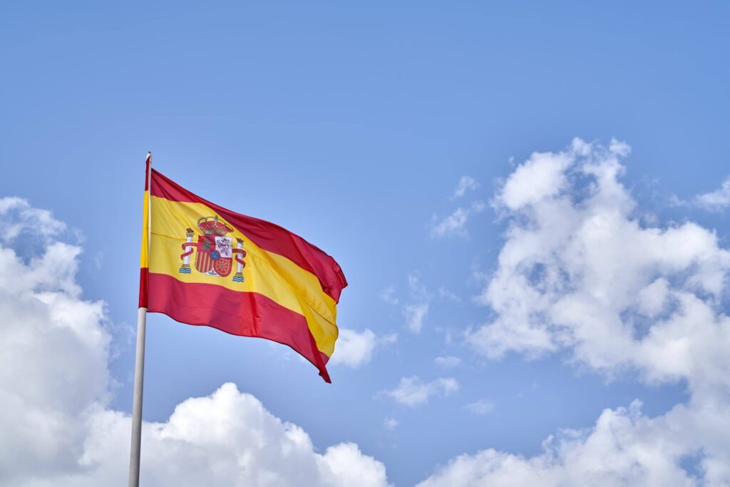 The red and yellow flag of Spain with the coat of arms, waving in the wind against a blue sky with white clouds.